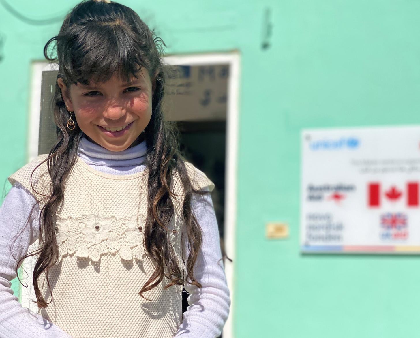 Kawthar, 10, smiles shyly at the camera as she stands outside in front of a green building.