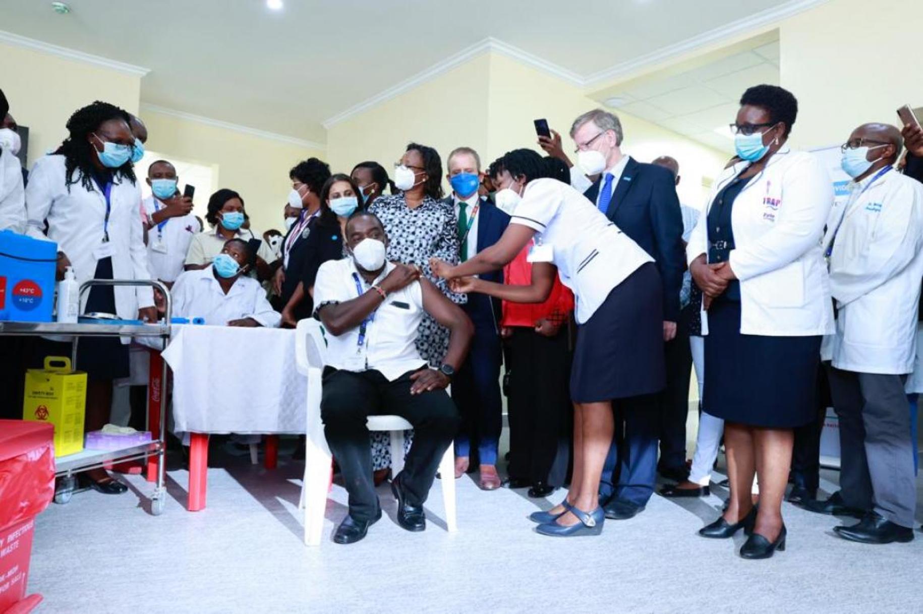A large group of people wearing masks stand around a seated man receiving a vaccination.. 