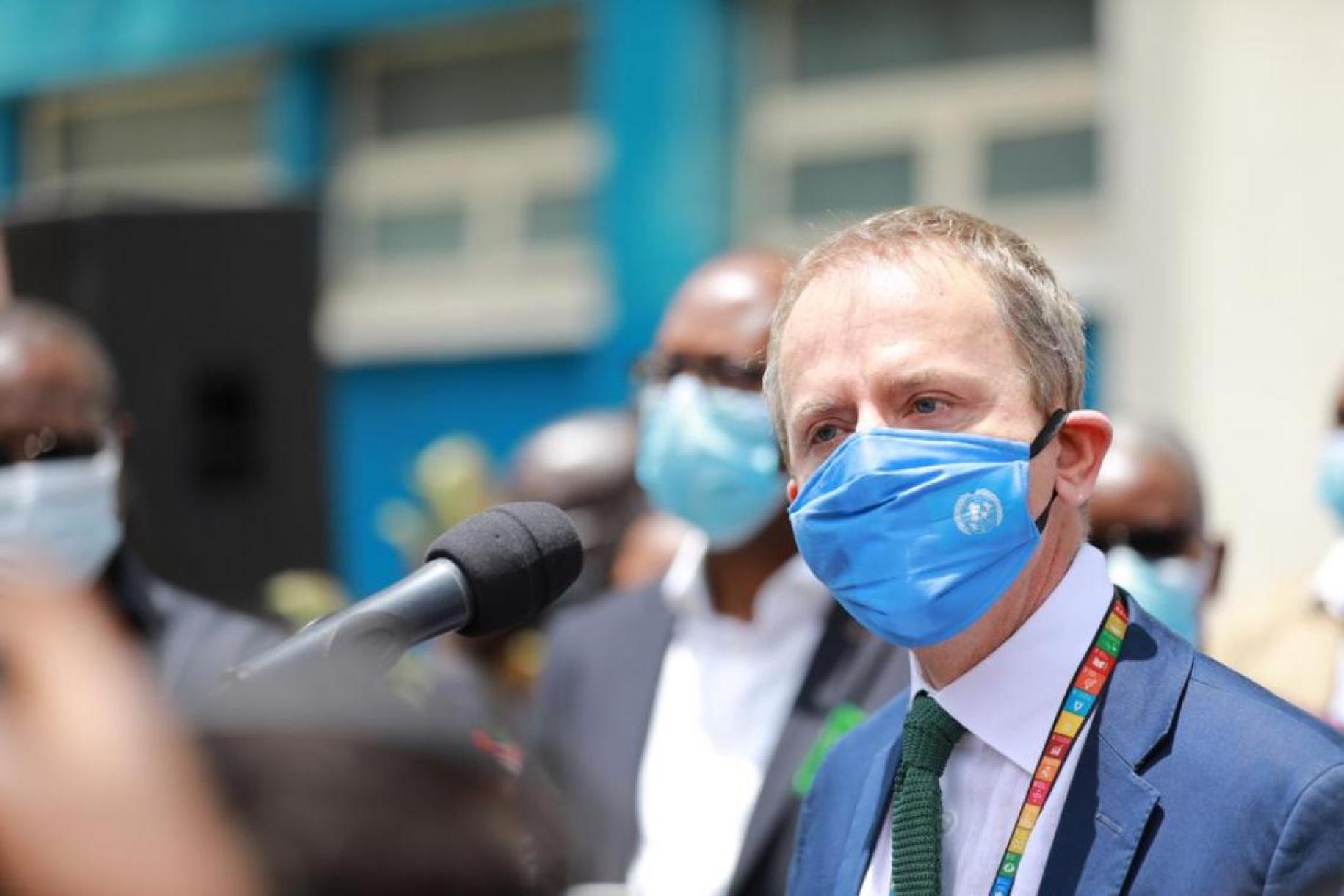People in masks stand behind a man with a light blue mask in a green tie and blue suit talking into a microphone. 