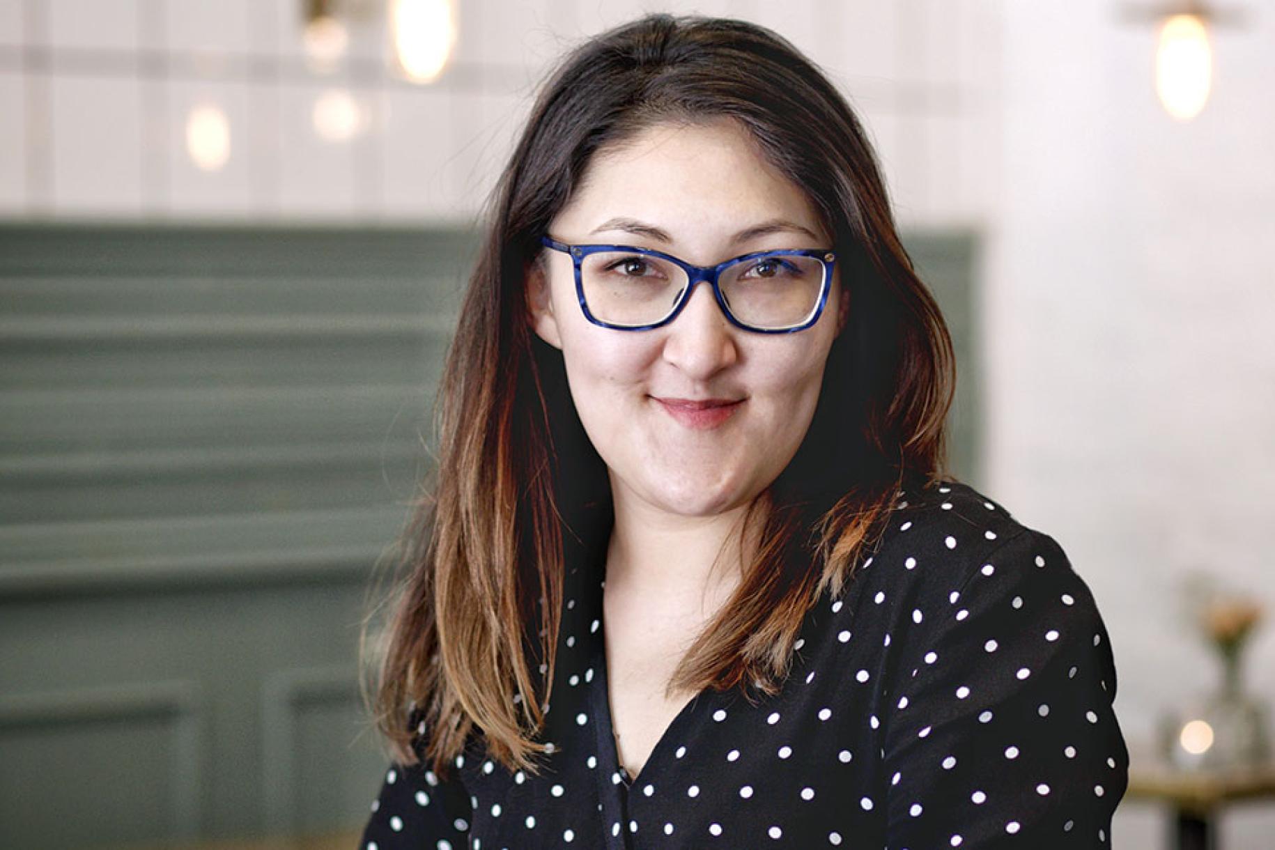 Une femme aux cheveux bruns et portant  des lunettes et une chemise noire à pois sourit à la caméra.