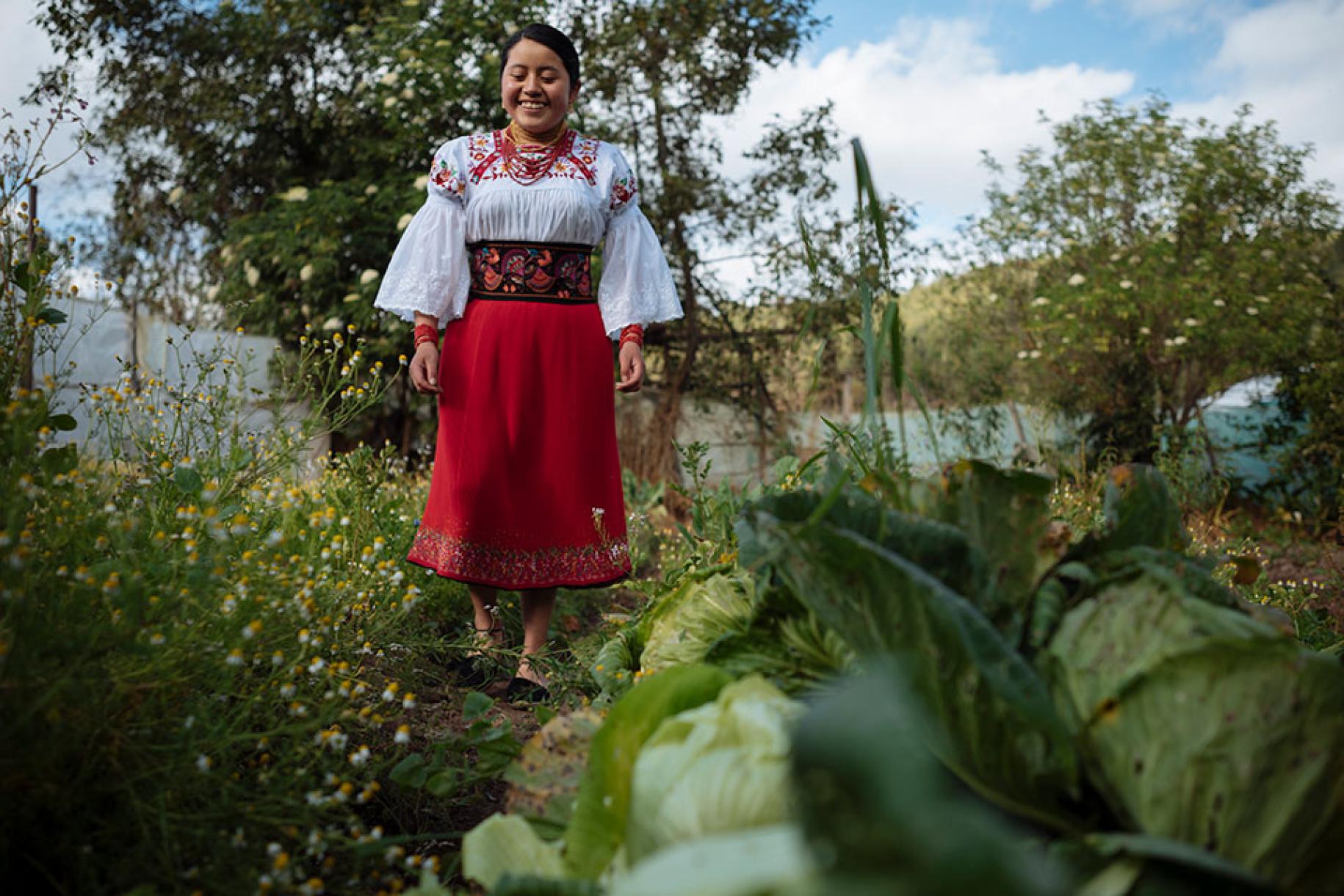 Une jeune fille souriante en costume traditionnel se tient dans un jardin entourée d'arbres et de fleurs.