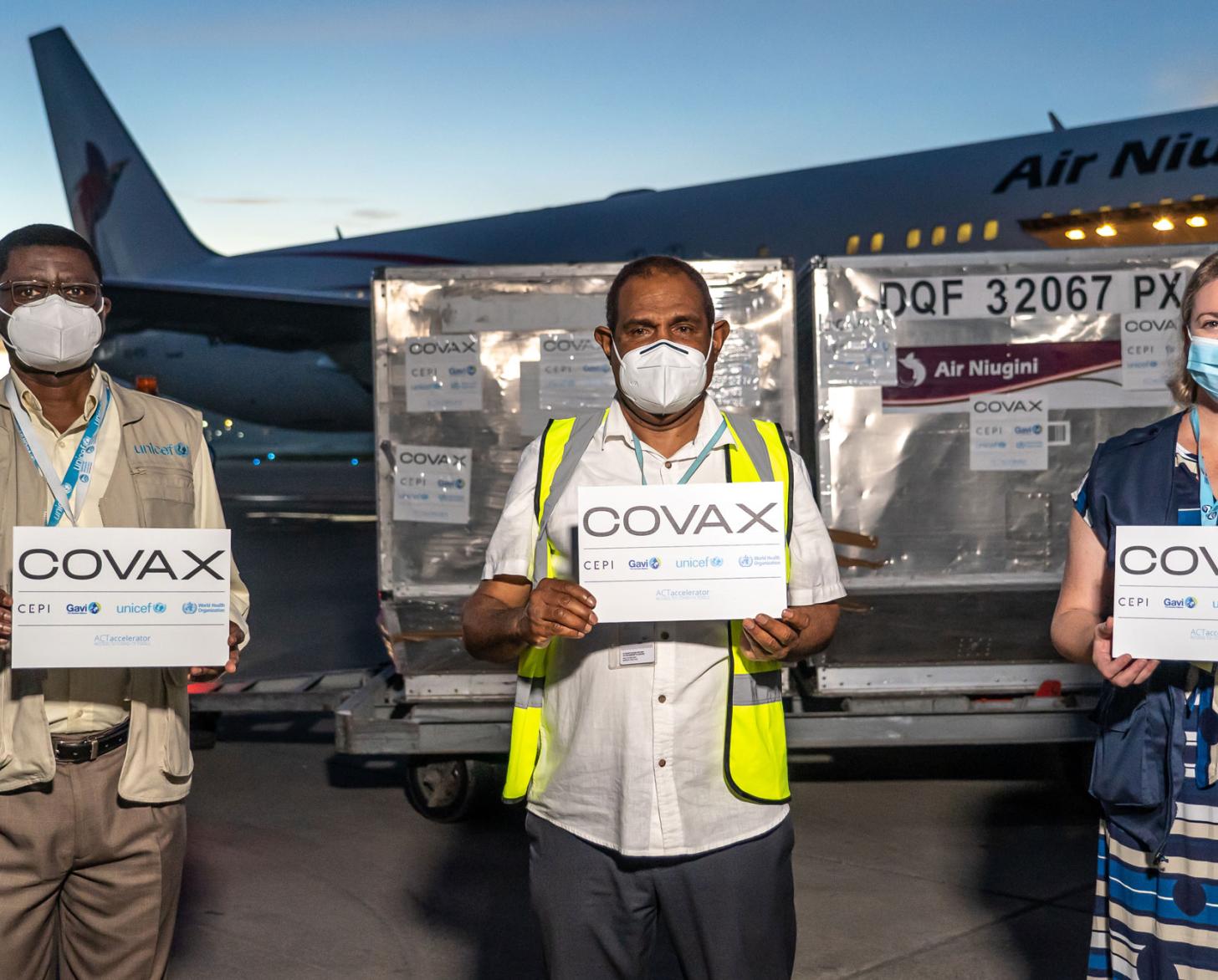 Three people in face masks hold COVAX signs stand in front of a delivery of the COVID-19 vaccines and an airplane.