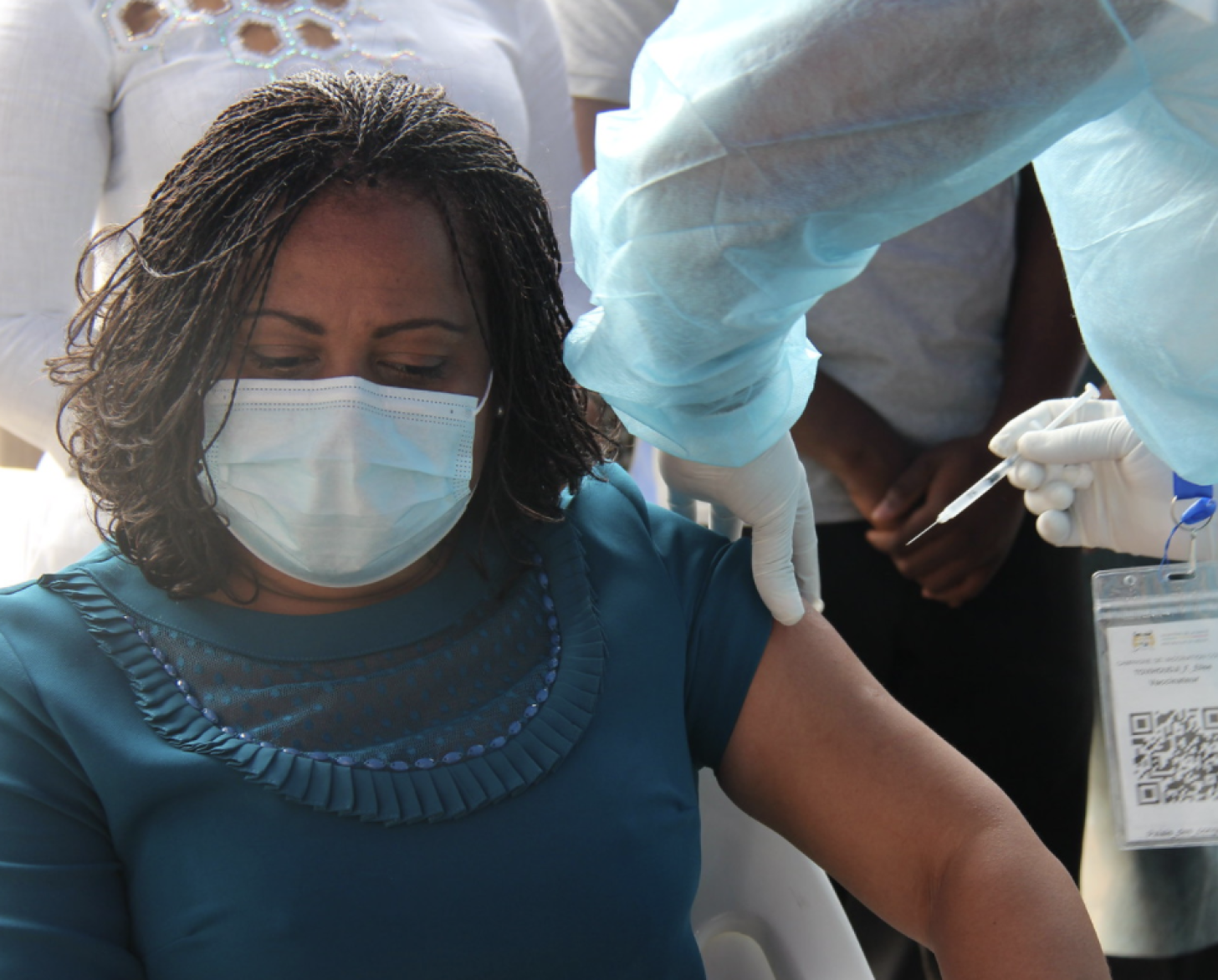 Una mujer vestida con una camisa verde azulado, mira hacia abajo mientras es vacunada por una persona con equipo de protección.