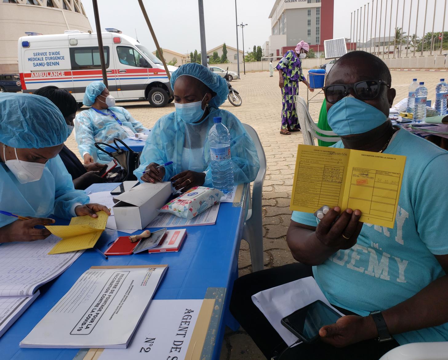 Un hombre con gafas de sol, una camisa azul y una mascarilla azul muestra su formulario de vacunación a la cámara mientras dos trabajadores de la salud se sientan cerca de él.