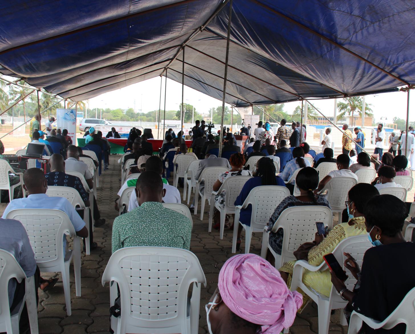 Muchas personas, manteniendo la distancia social recomendada entre si, se sientan bajo una carpa al aire libre, de espaldas a la cámara.