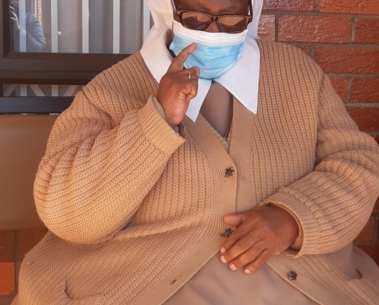 A woman in a tan sweater and dress holds up one hand while the other hand rests on her lap.