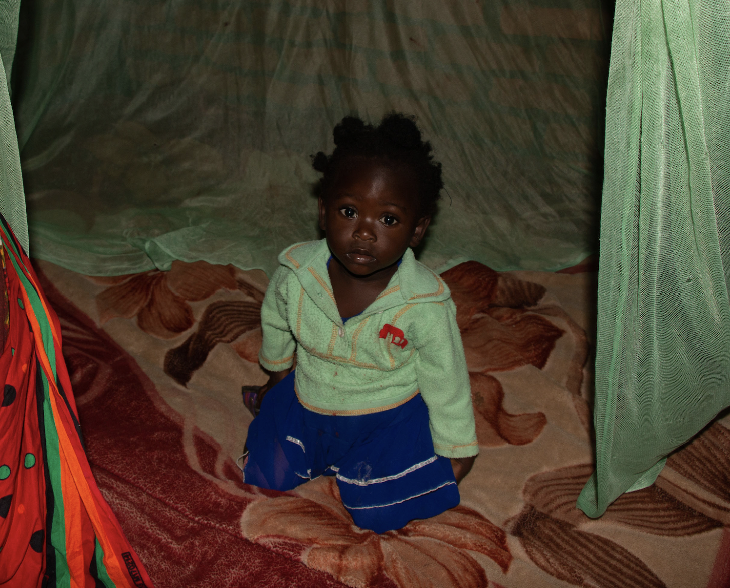 A young girl in a green sweater and shorts sits under a green mosquito net.