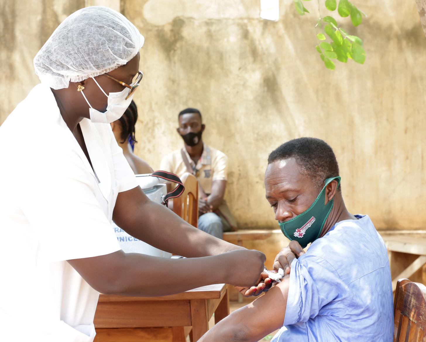 Un hombre con una mascarilla verde recibe la vacuna contra la COVID-19 de una mujer vestida con equipo médico. 