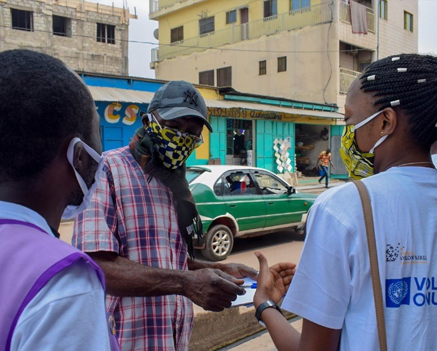Three people in masks stand on the side of the road as a green car passes by behind them.