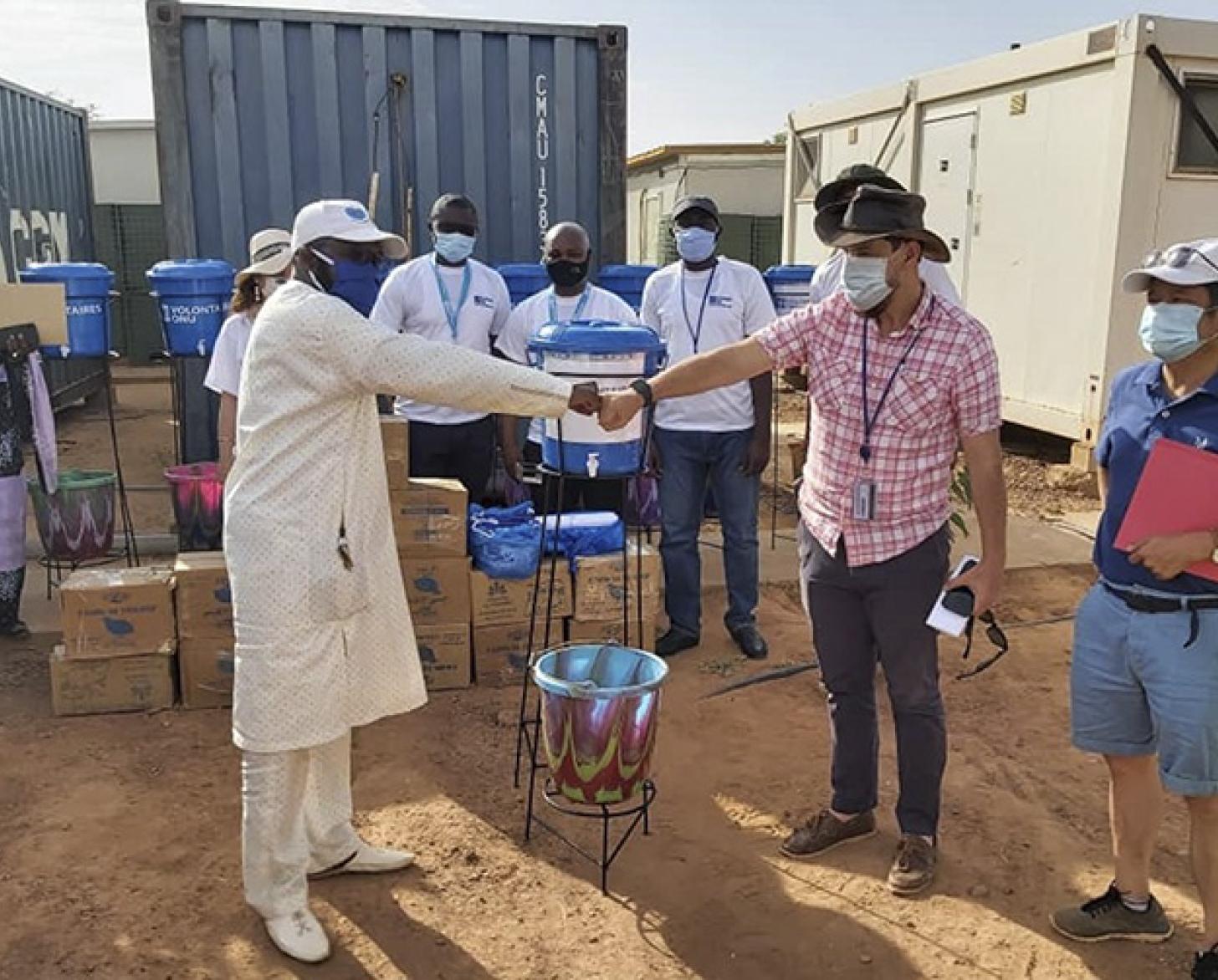 Two men fist bump their hands while several other people stand around them near large containers.