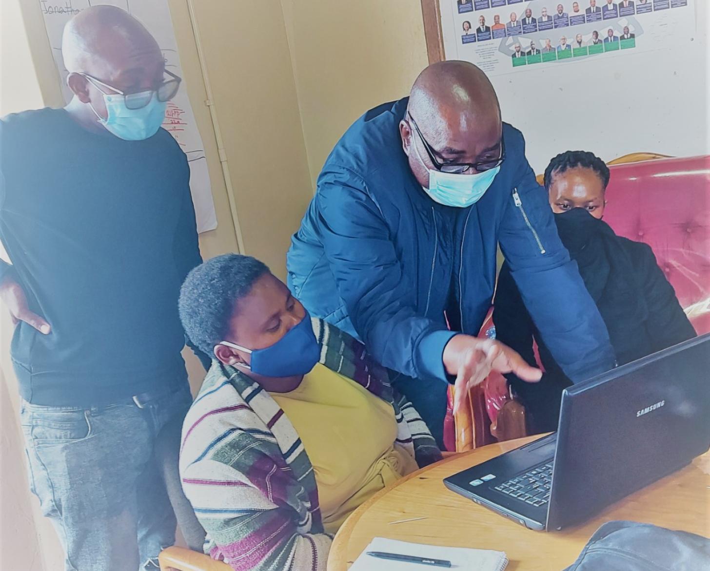 Two men stand over two people sitting by a computer with his hand stretched out.