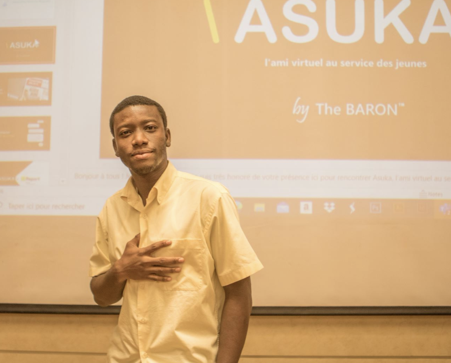 Un joven, con camisa blanca abotonada, se sitúa frente a un proyector llevándose la mano al corazón. 