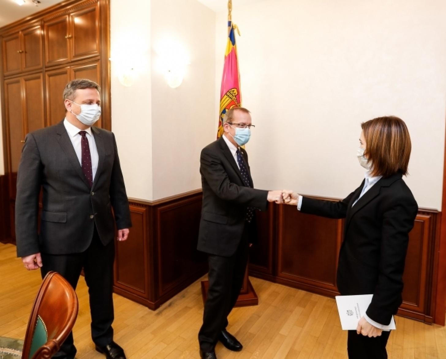 Un hombre y una mujer trajeados se dan la mano mientras otro hombre se sitúa junto a ellos.