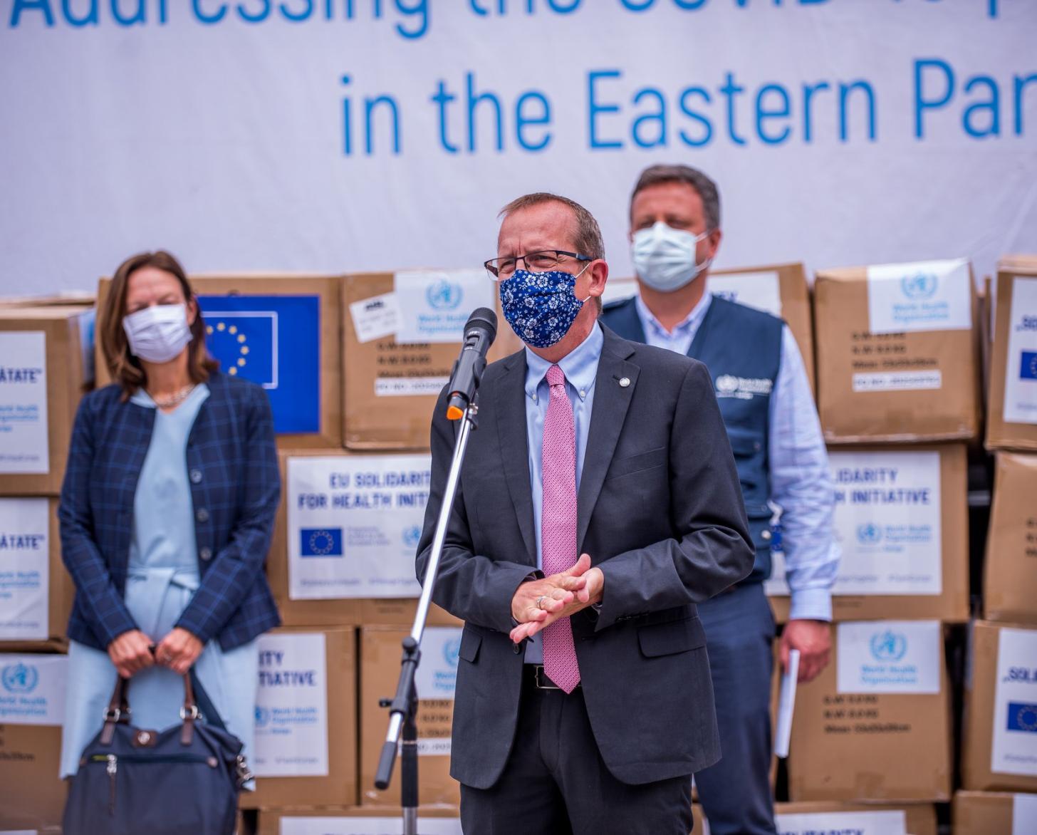 Cuatro personas con mascarillas se sitúan delante de las cajas de vacunas, asociadas al mecanismo COVAX, mientras un hombre habla con un micrófono. 
