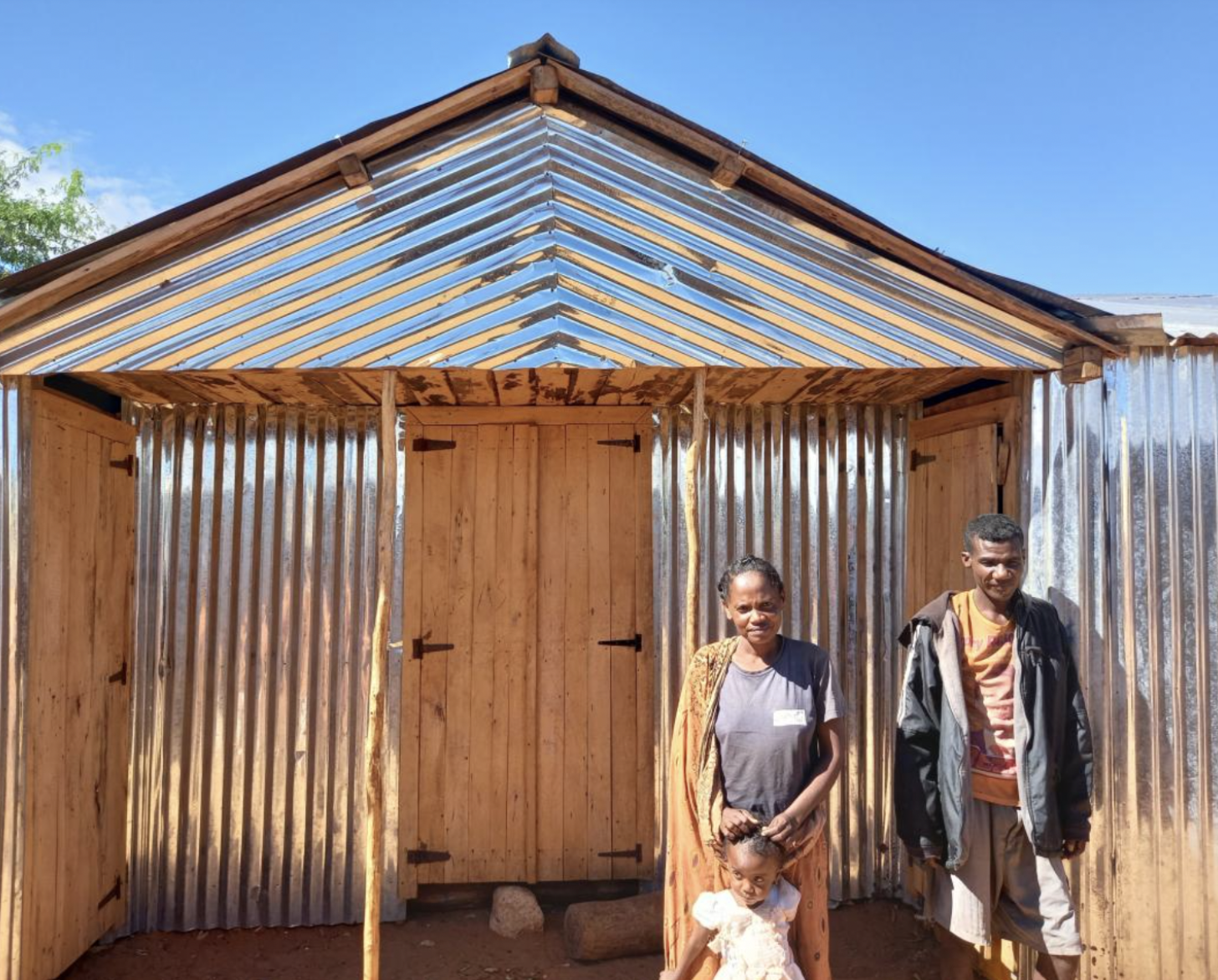 The image shows Fagnosea and Masy with their child standing in front of the house they built.