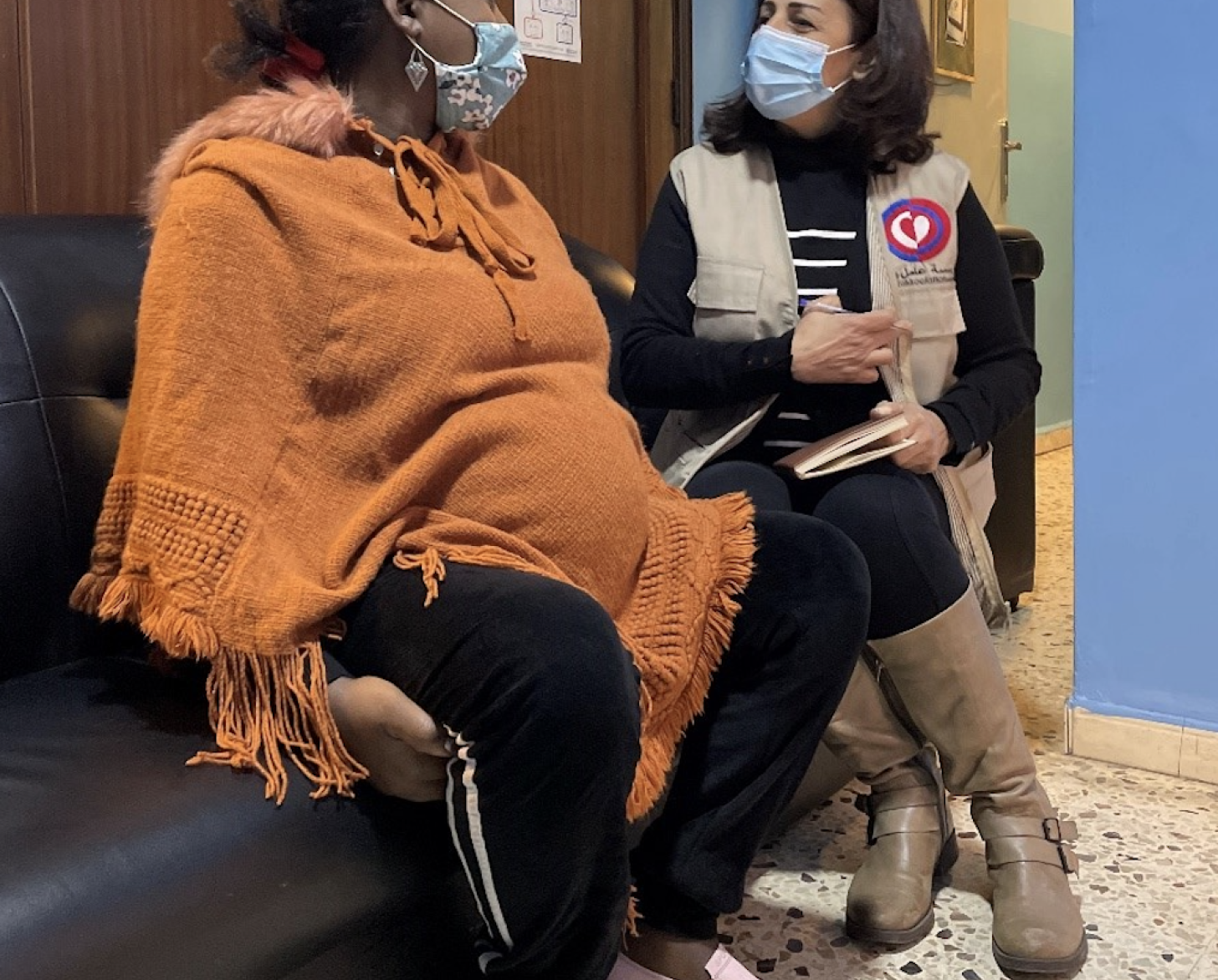 Dos mujeres, una con jersey naranja y otra con camisa negra y chaleco terracota, se sientan en un sofá a hablar entre ellas. 