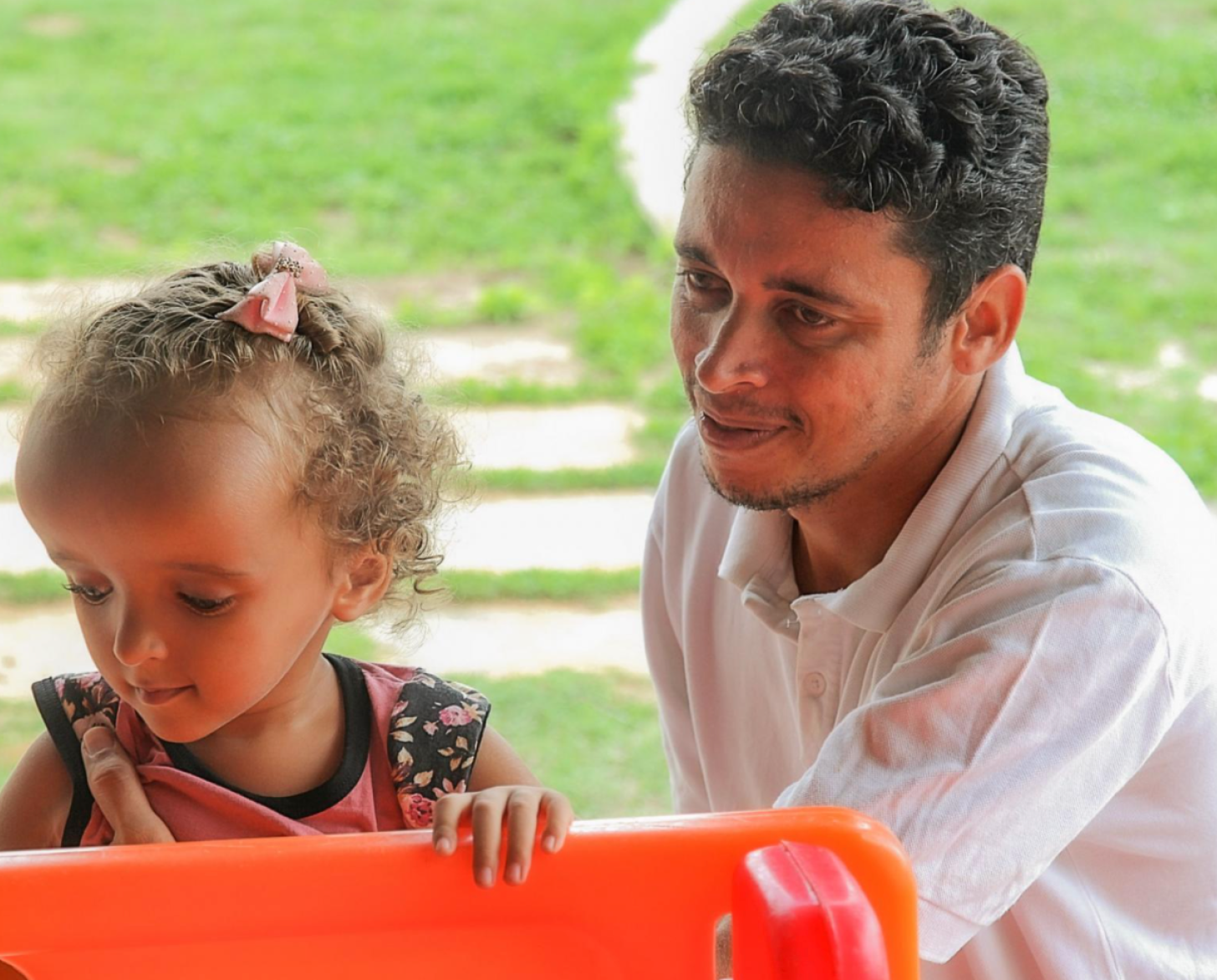 A man and a child look at an orange toy together in an outdoor space.