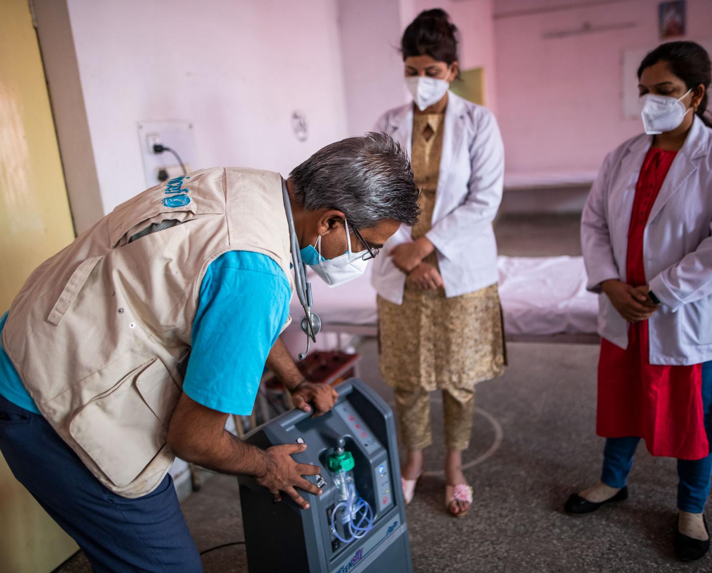 A man, with a UNICEF vest on, with an oxygen concentrator shows two medical professionals how it works. 