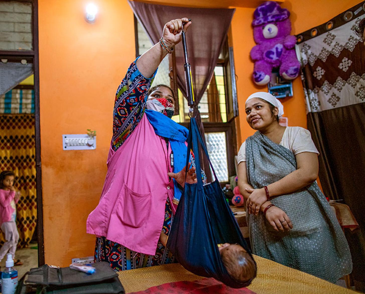 Two women weigh a baby inside a home. 