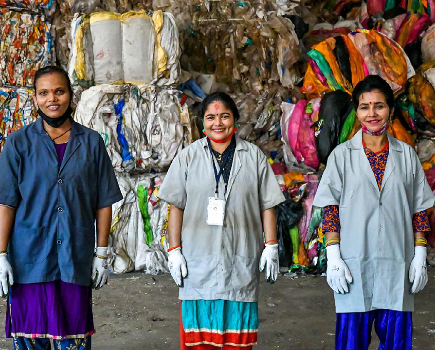 Three women smile at the camera in front of large piles of packaged waste.