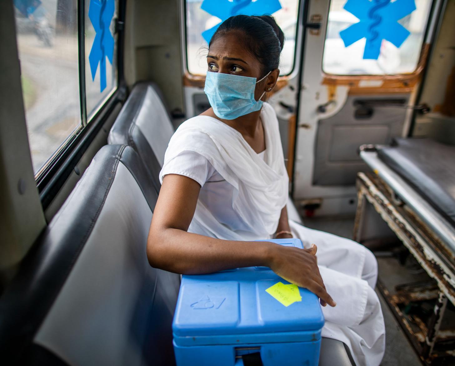 Dans l'ouest de l'Inde, une sage-femme infirmière portant un masque de protection respiratoire est assise dans une ambulance, la main posée sur une glacière contenant des vaccins anti COVID-19, et lance un regard inquiet à travers la fenêtre.