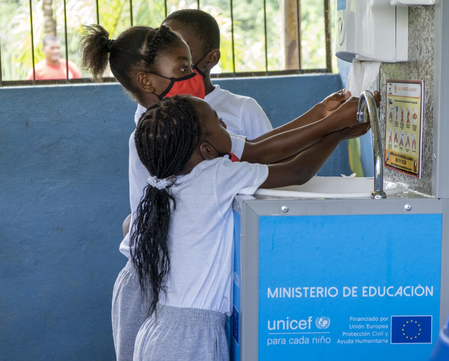 Trois jeunes enfants se lavent les mains dans une station de lavage des mains installée dans leur école.