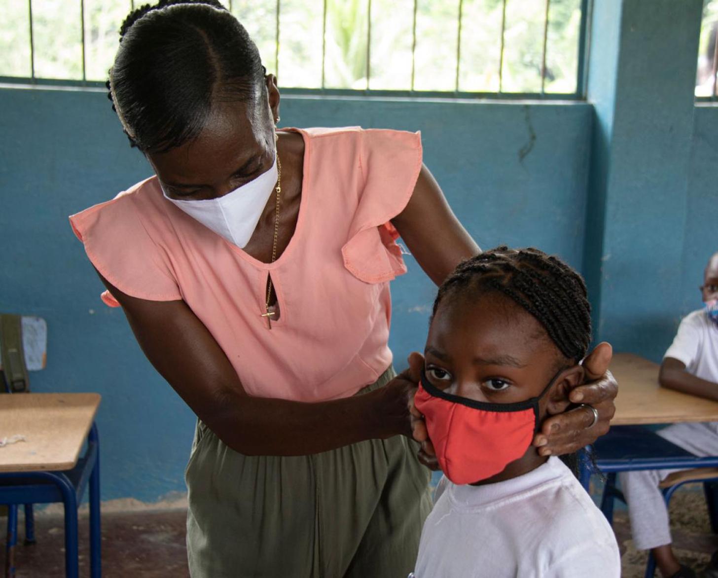 Gros plan sur une enseignante de l’enseignement primaire portant un masque de protection respiratoire qui aide un élève à ajuster son masque sur le visage dans une salle de classe.