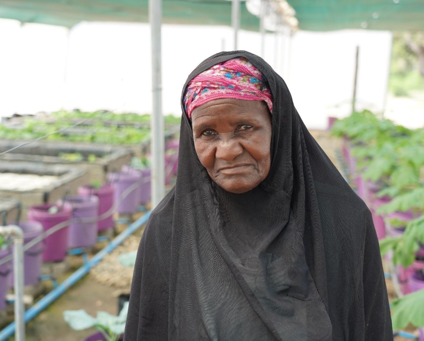 A woman in a black veil looks at the camera, and stands in between a row of plants in a greenhouse.