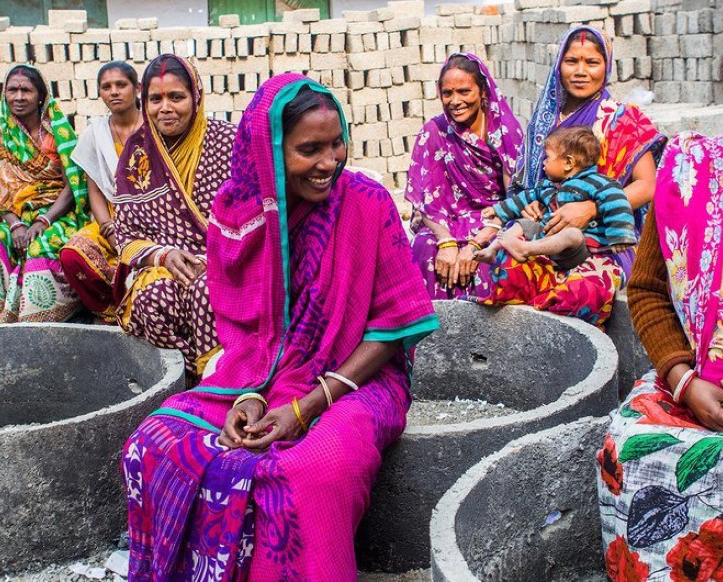 Mujeres vestidas con saris de colores se sientan y hablan, con un fondo de ladrillos para una futura estructura detrás de ellas.
