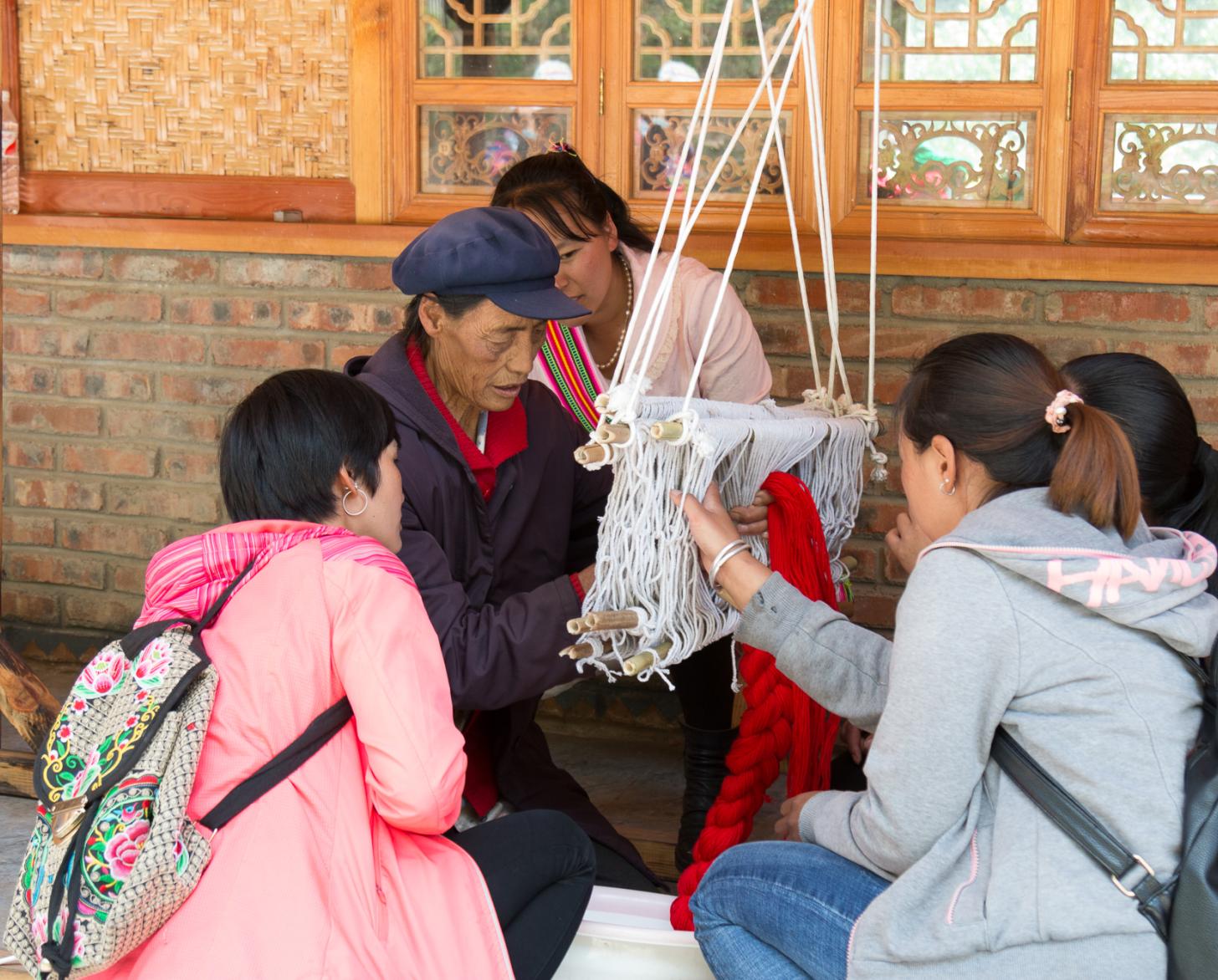 4 شابات يجلسن حول امرأة عجوز يشاهدنها وهي تنسج الصوف.