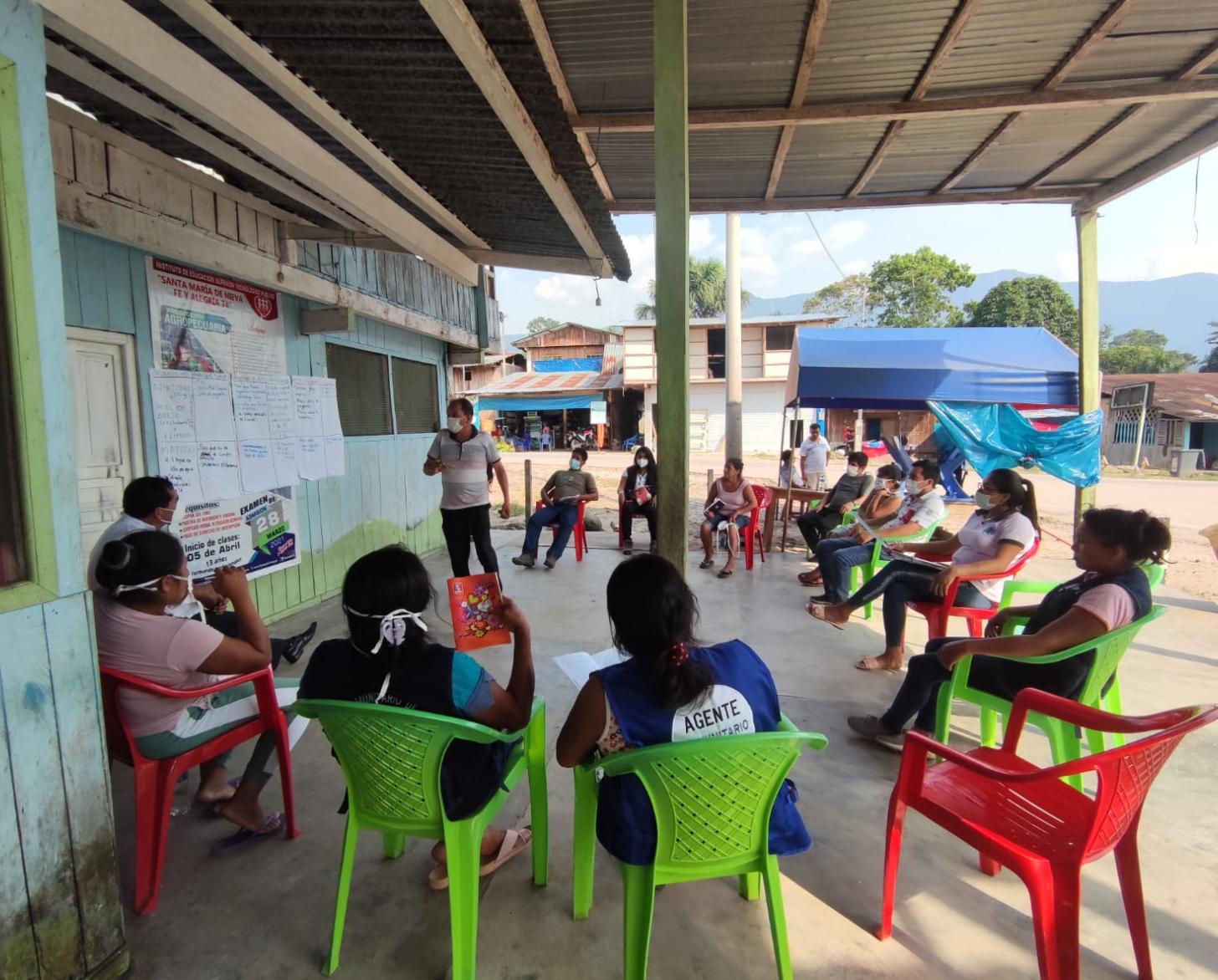 Un grupo de personas sentadas en sillas de plástica a manera de círculo, en un espacio exterior semi techado.