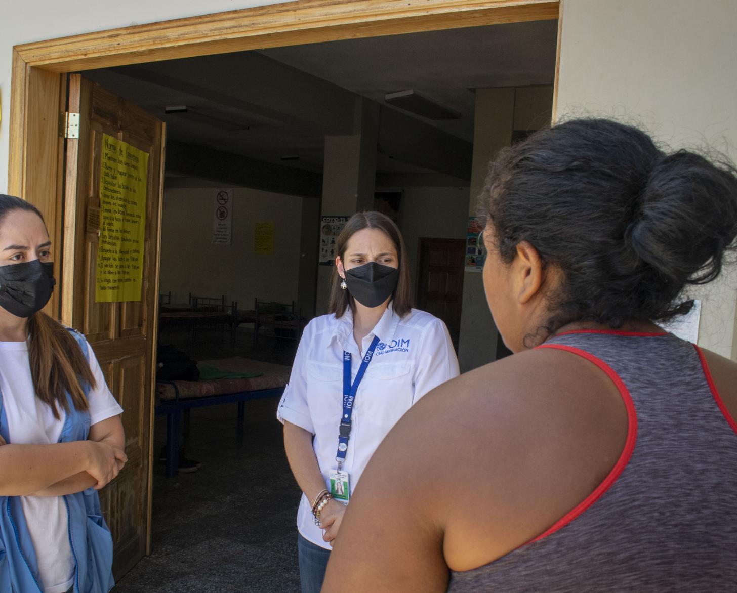 Una mujer, de espaldas a la cámara, habla con dos personas con mascarillas, una de ellas con un chaleco que la identifica como miembro del sistema de las Naciones Unidas y la otra con un distintivo de la ONU.