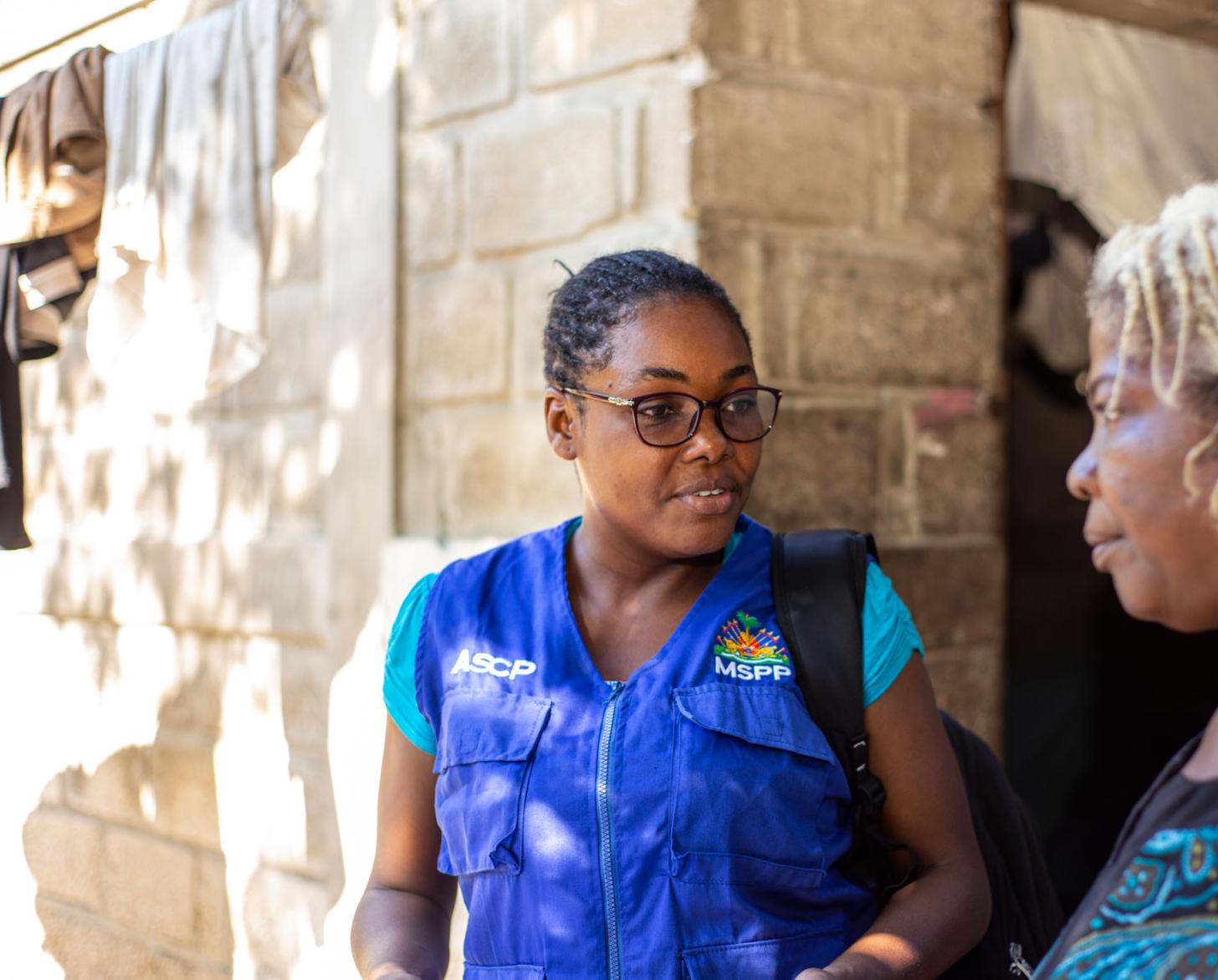 Gros plan sur une femme portant un uniforme bleu et des lunettes et discutant avec une autre femme aux cheveux décolorés.