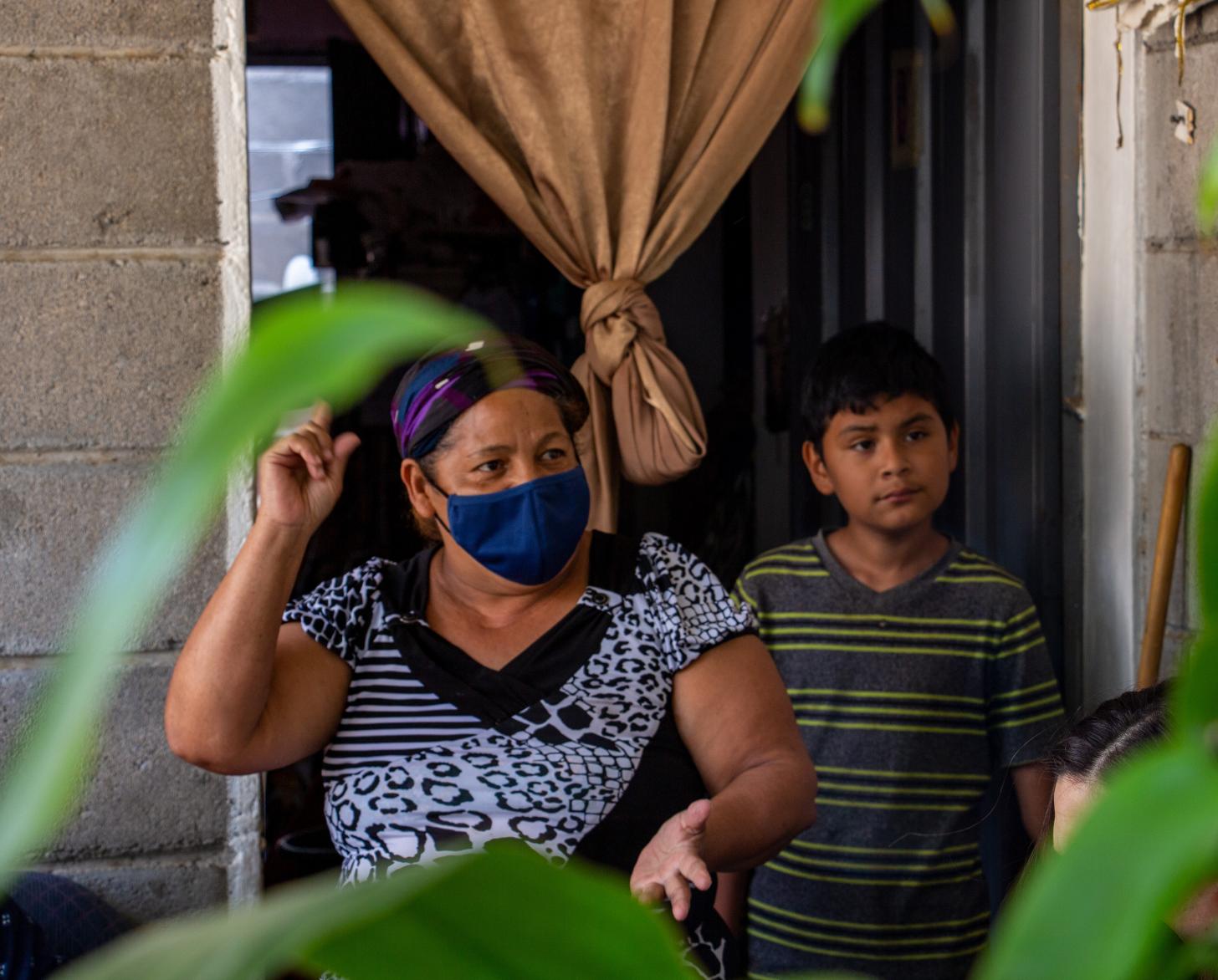 Delante de la puerta de una casa, una mujer con mascarilla y un niño hablan con una persona que está fuera de la toma.
