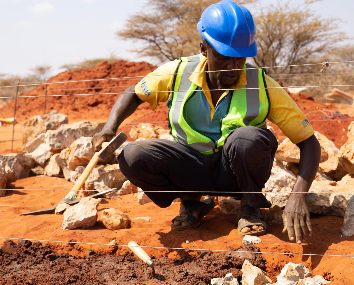 Man in a green vest and a blue hard hat kneels on the ground with tools
