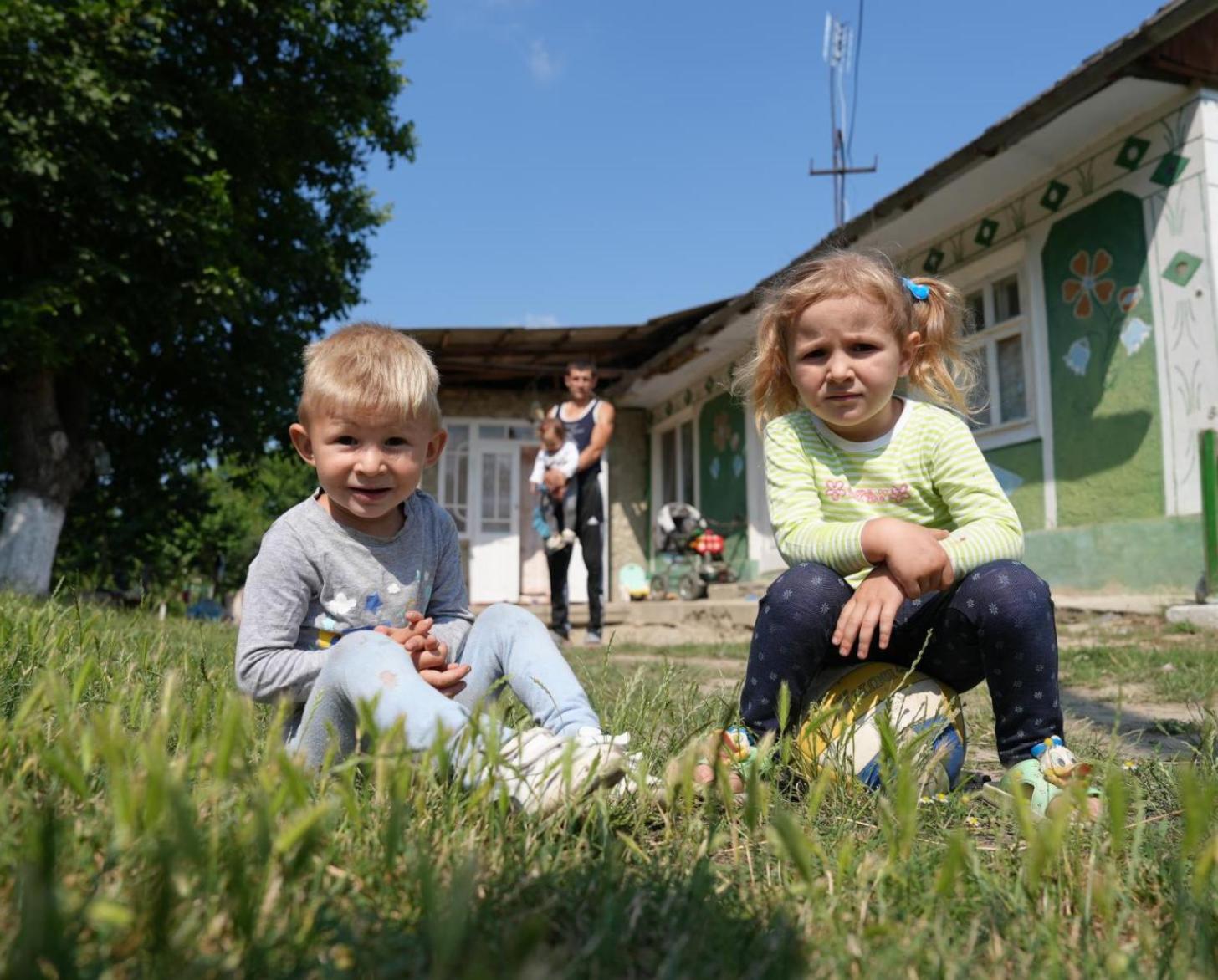 Two children sit on grass in front of a house and smile.