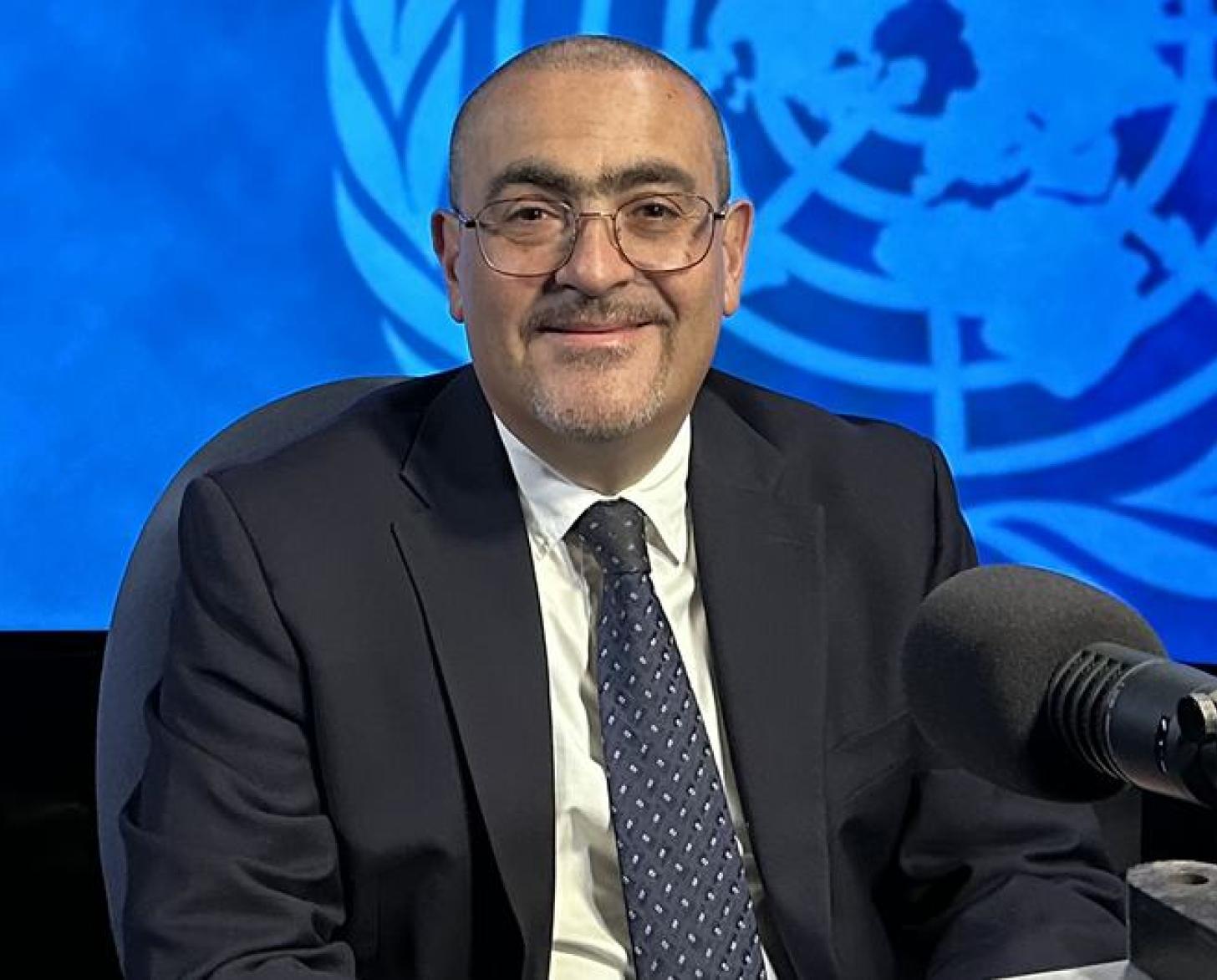 Мужчина в очках (Рамиз Алакбаров) улыбается на камеру, сидя перед микрофоном.