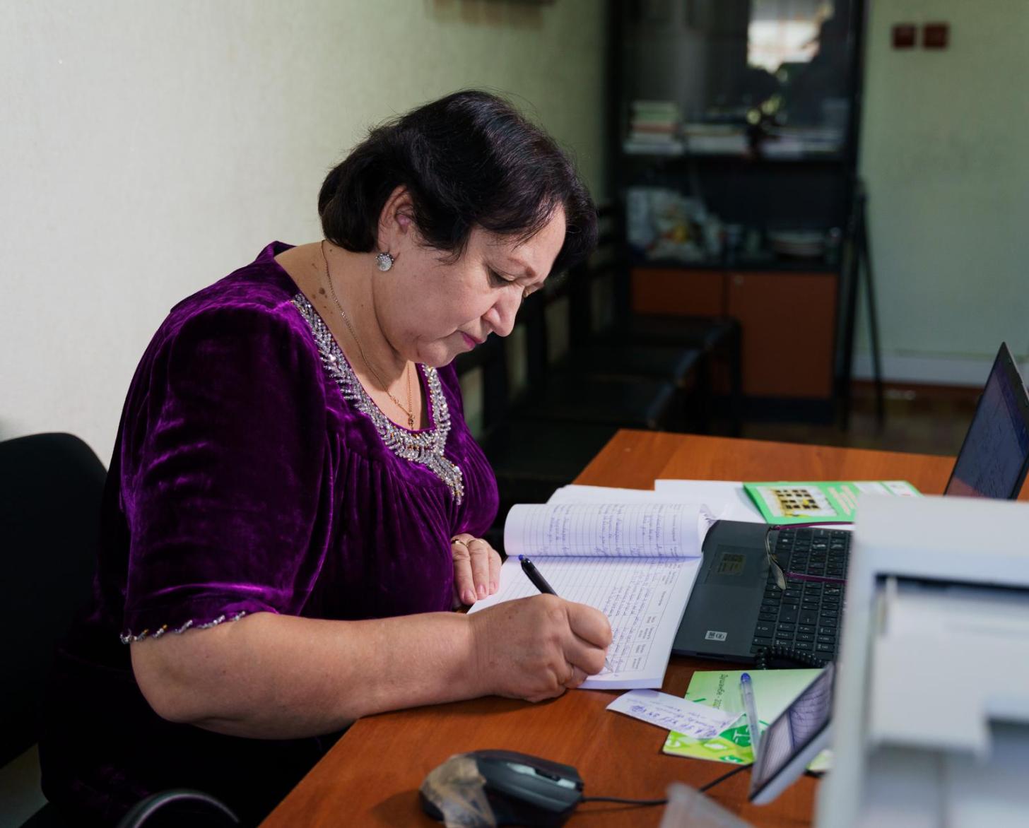 A woman in a purple dress sits at a desk writing in a notebook, papers strewn all around her.