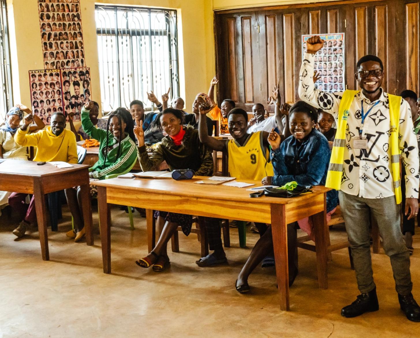 A man in a yellow and white shirt raises his hand in joy as he stands in front of a classroom full of students at desks who are also doing the same