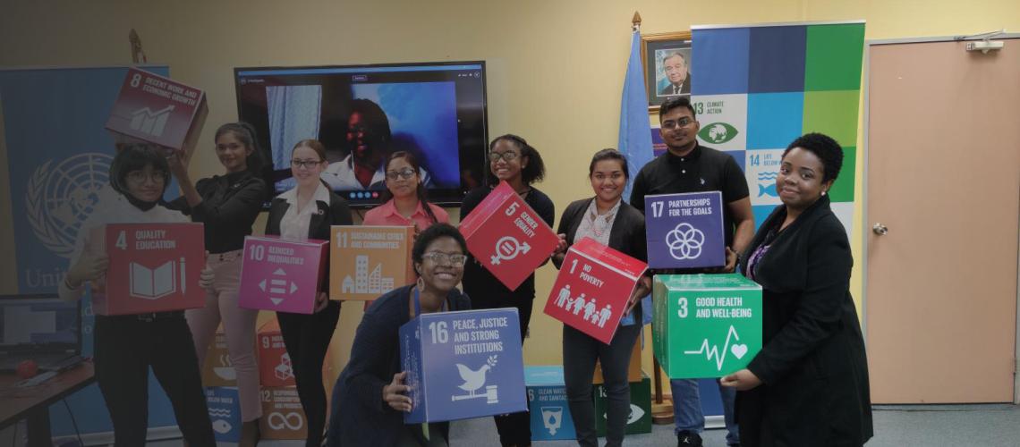 Young people in a room stand in front of a video call and smile at the camera as they hold blocks decorated with SDG logos.